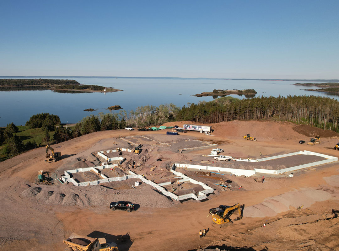 Aerial construction photograph of the new St. Anne Centre site in Arichat, with foundations being poured and the Isle Madame coastline in the background